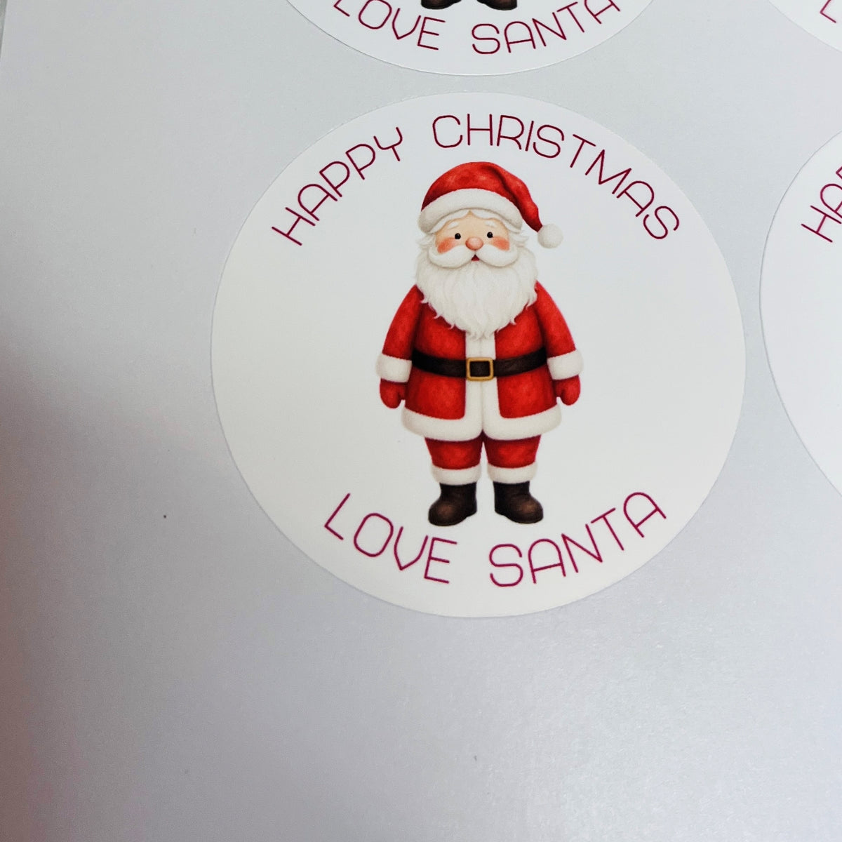 A round white sticker featuring a cartoon Santa Claus in a red suit with a white beard, surrounded by the text 'HAPPY CHRISTMAS LOVE SANTA' in red, shown on a white background.