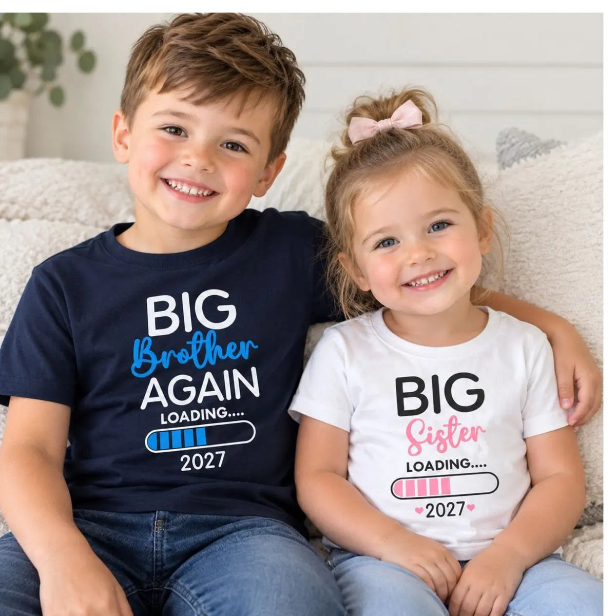A smiling young boy and girl sit together on a light sofa wearing matching graphic T-shirts. The boy wears a navy T-shirt with white and blue text reading 'BIG Brother AGAIN LOADING, and the young girl wears a white T-shirt with the graphic 'Big Sister Loading'