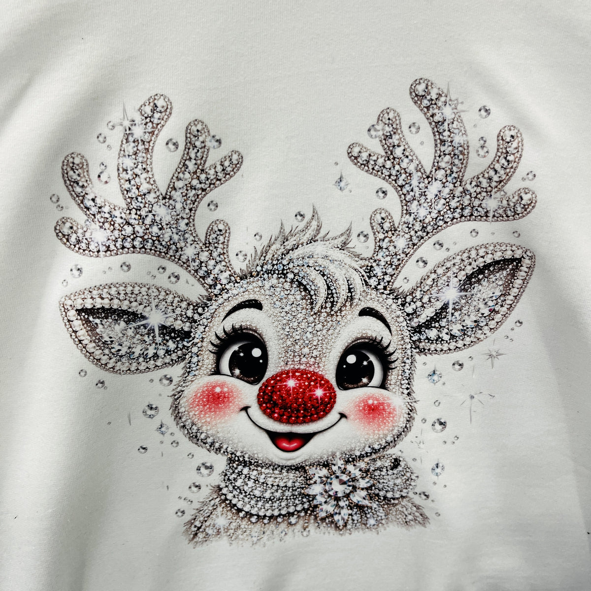 A close-up of a white garment featuring a rhinestone-embellished cartoon reindeer face with large black eyes, pink cheeks, a red nose, and silver antlers.
