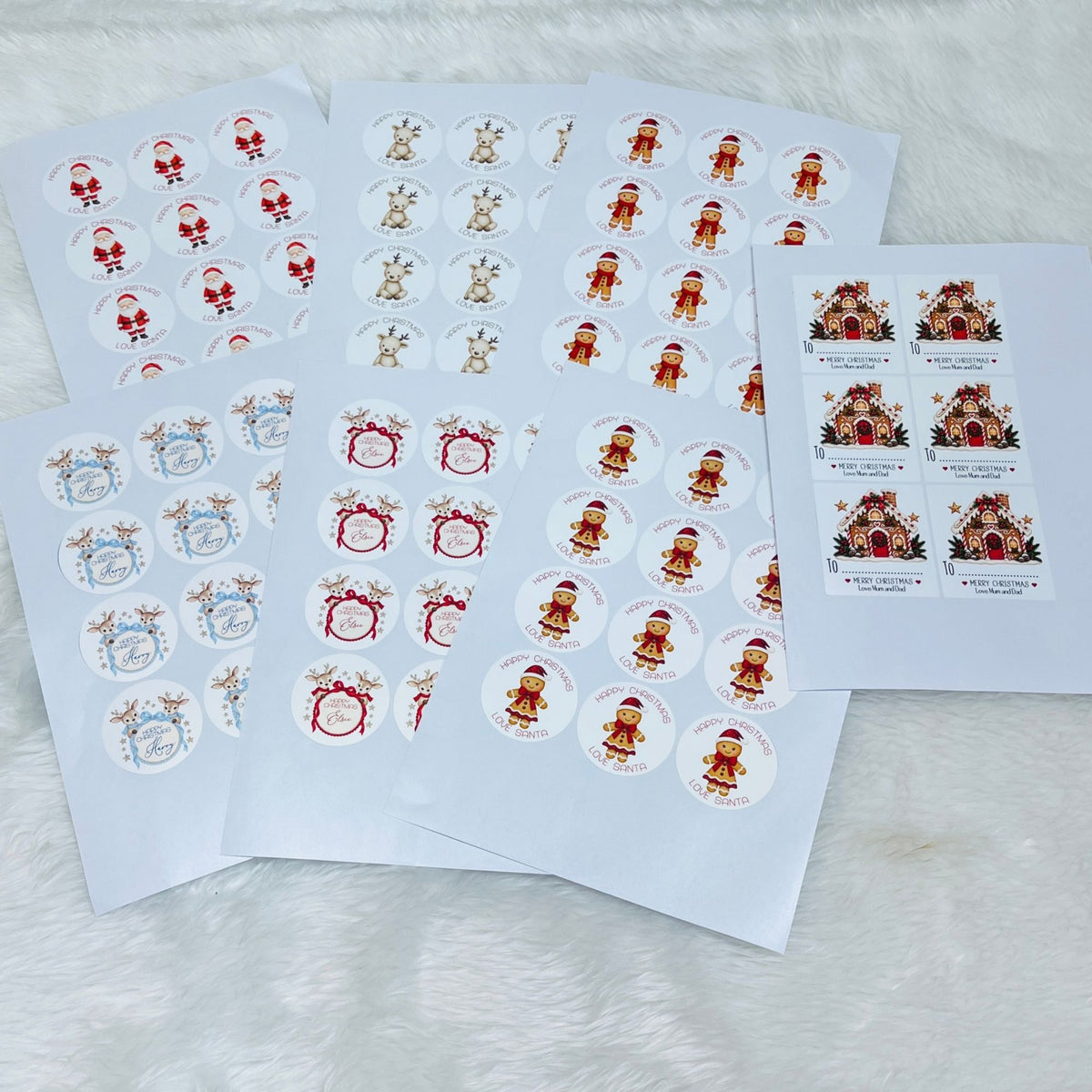 Several sheets of round Christmas-themed stickers laid out on a white faux-fur surface. The stickers feature Santa Claus, reindeer, holiday wreaths, and gingerbread house designs on white backing sheets.