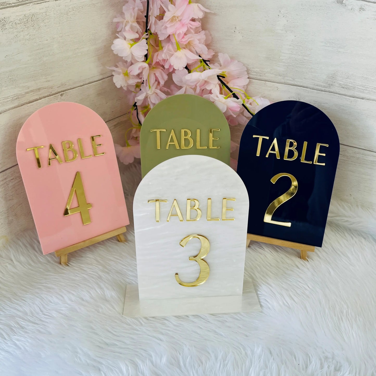 Four table number holders in different colors with gold text on a soft white background.