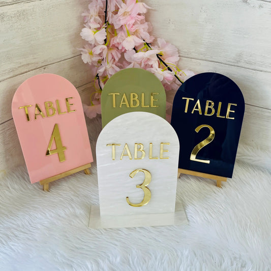 Four table number holders in different colors with gold text on a soft white background.