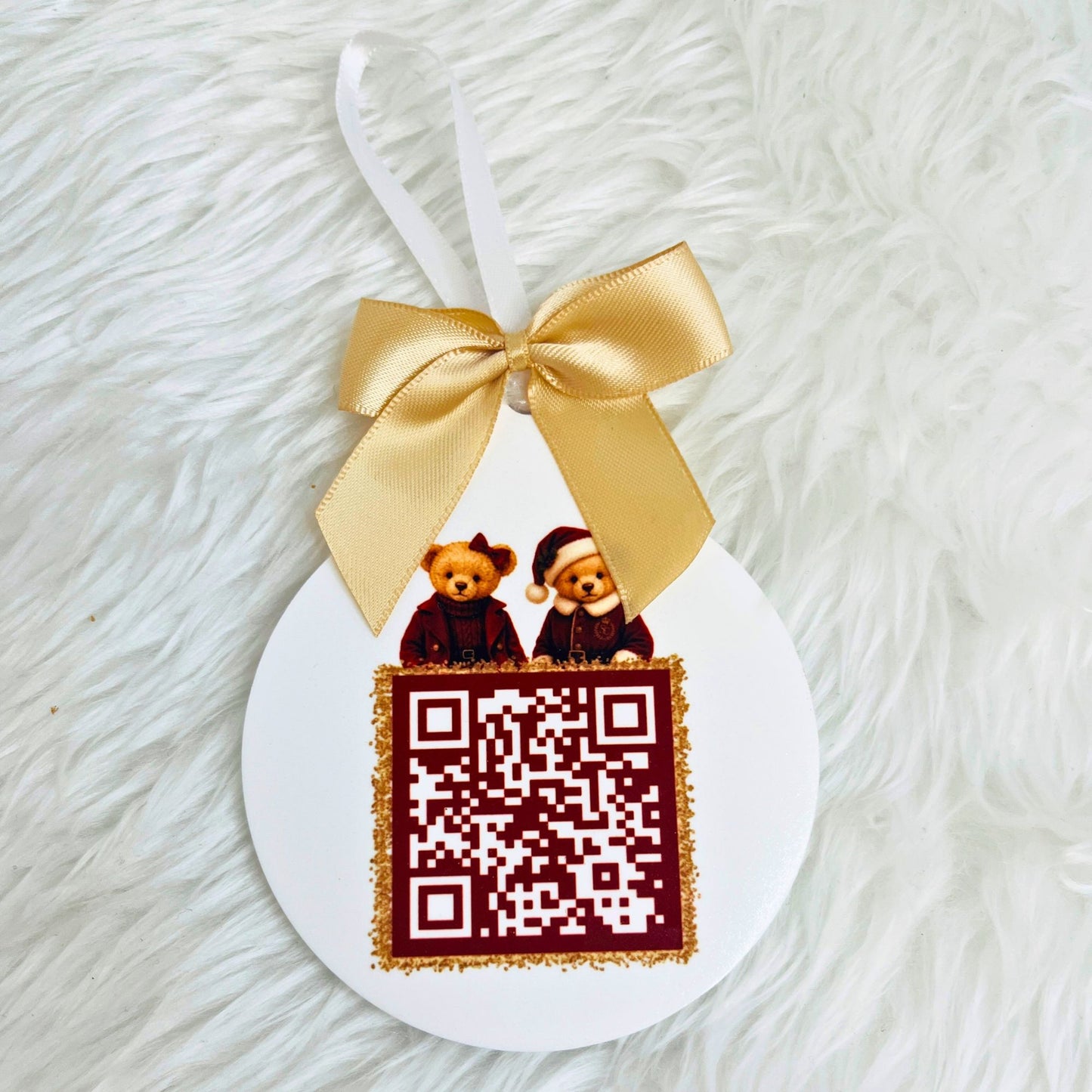 A round white hanging ornament with a white ribbon loop, decorated with a large gold satin bow, a printed santa tracker QR code with gold glitter trim, and two small teddy bears wearing Santa hats above the code. The ornament is photographed on a white faux-fur surface.