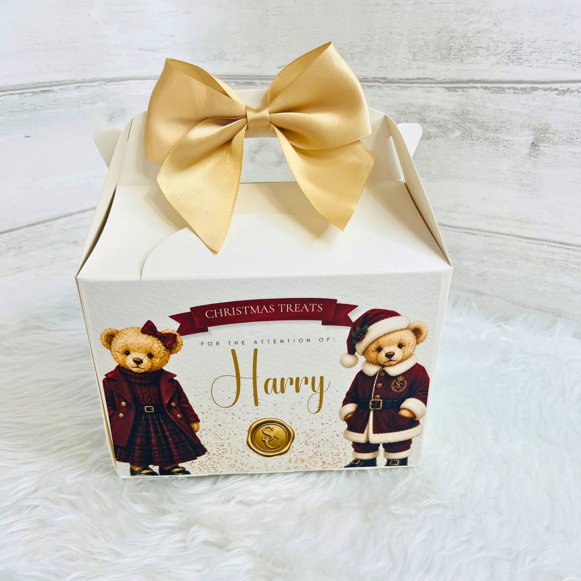 A white square gift box for Christmas treats, decorated with a large gold satin bow on top. The front of the box features illustrations of two teddy bears in holiday clothing and the text 'CHRISTMAS TREATS FOR THE ATTENTION OF Harry'.