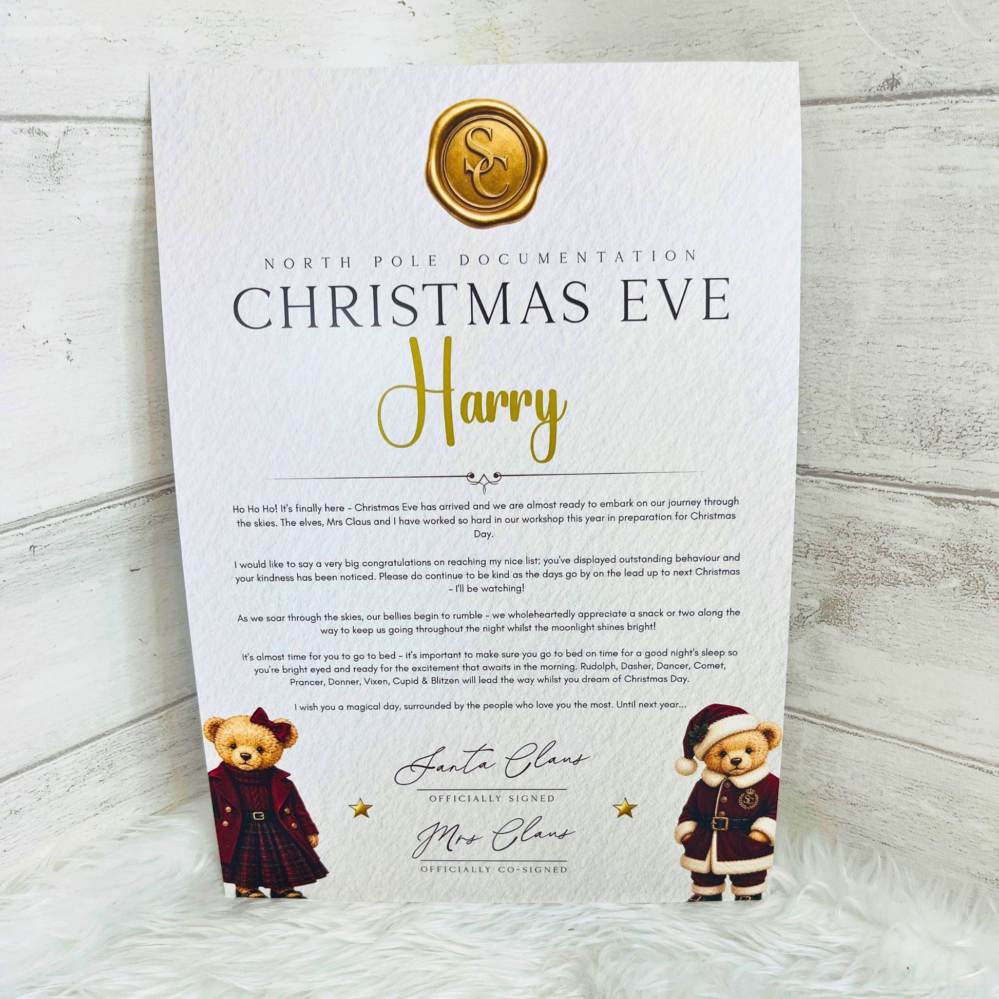 A printed Christmas Eve North Pole Documentation letter for 'Harry' is displayed on a white faux fur surface against a light wood background. The letter features a gold foil seal at the top, a body of text, and illustrations of teddy bears dressed in Santa claus outfits at the bottom.