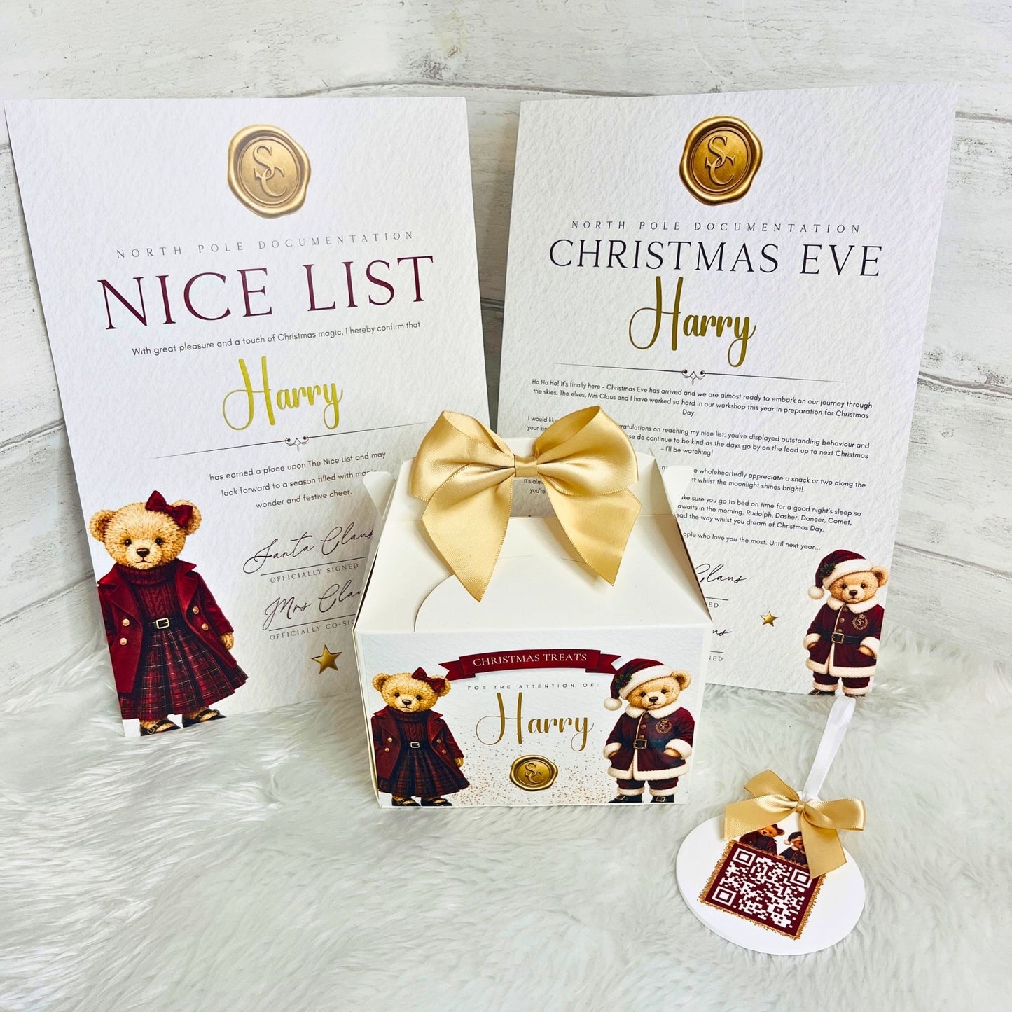 A white personalised Christmas gift box with a gold ribbon bow, displayed alongside matching printed cards and a small hanging ornament. The box and cards feature teddy bear illustrations dressed in holiday clothing and the names 'Harry' and 'Harry' printed on them.