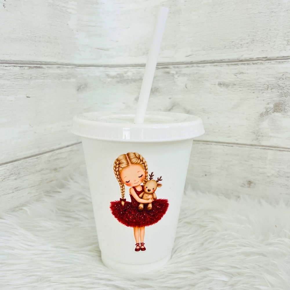 A white 16oz plastic cold cup tumbler with a lid and straw, featuring a cute illustration of a girl with braided hair in a red tutu glittery dress hugging a small reindeer, set against a rustic white wood and faux fur background.