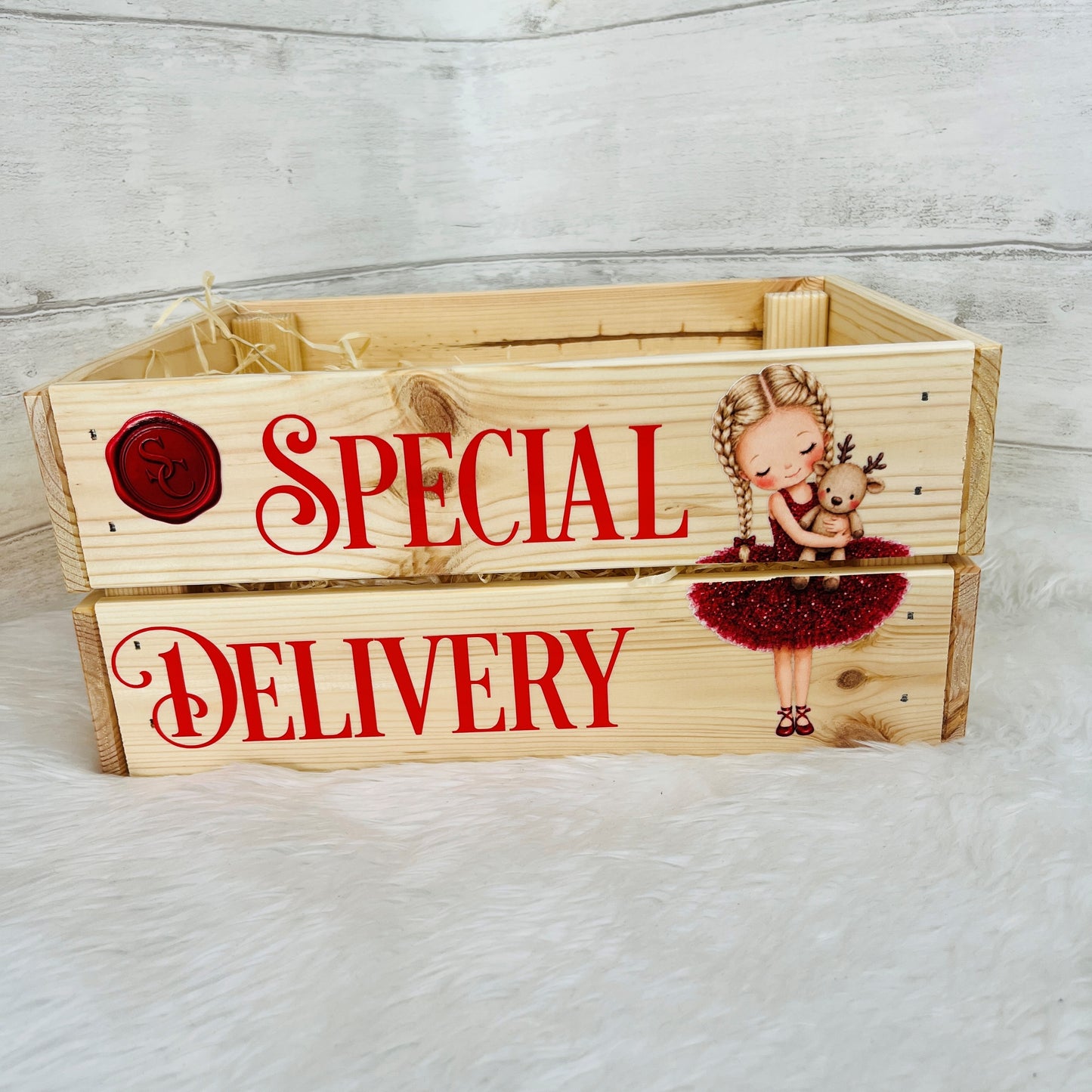 A light natural wooden crate  printed with red text reading 'SPECIAL DELIVERY' and a small illustration of a girl in a red dress hugging a reindeer, set on a white faux-fur surface against a pale wood plank backdrop.