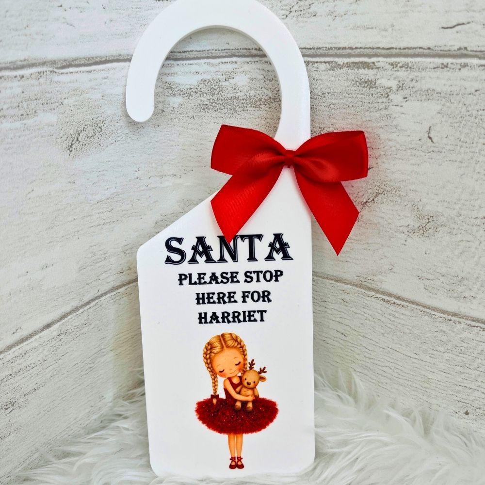 A white Christmas door hanger with a red ribbon bow, featuring the text 'SANTA PLEASE STOP HERE FOR HARRIET' and an illustration of a girl in a red tutu holding a reindeer, set against a rustic white wood background.
