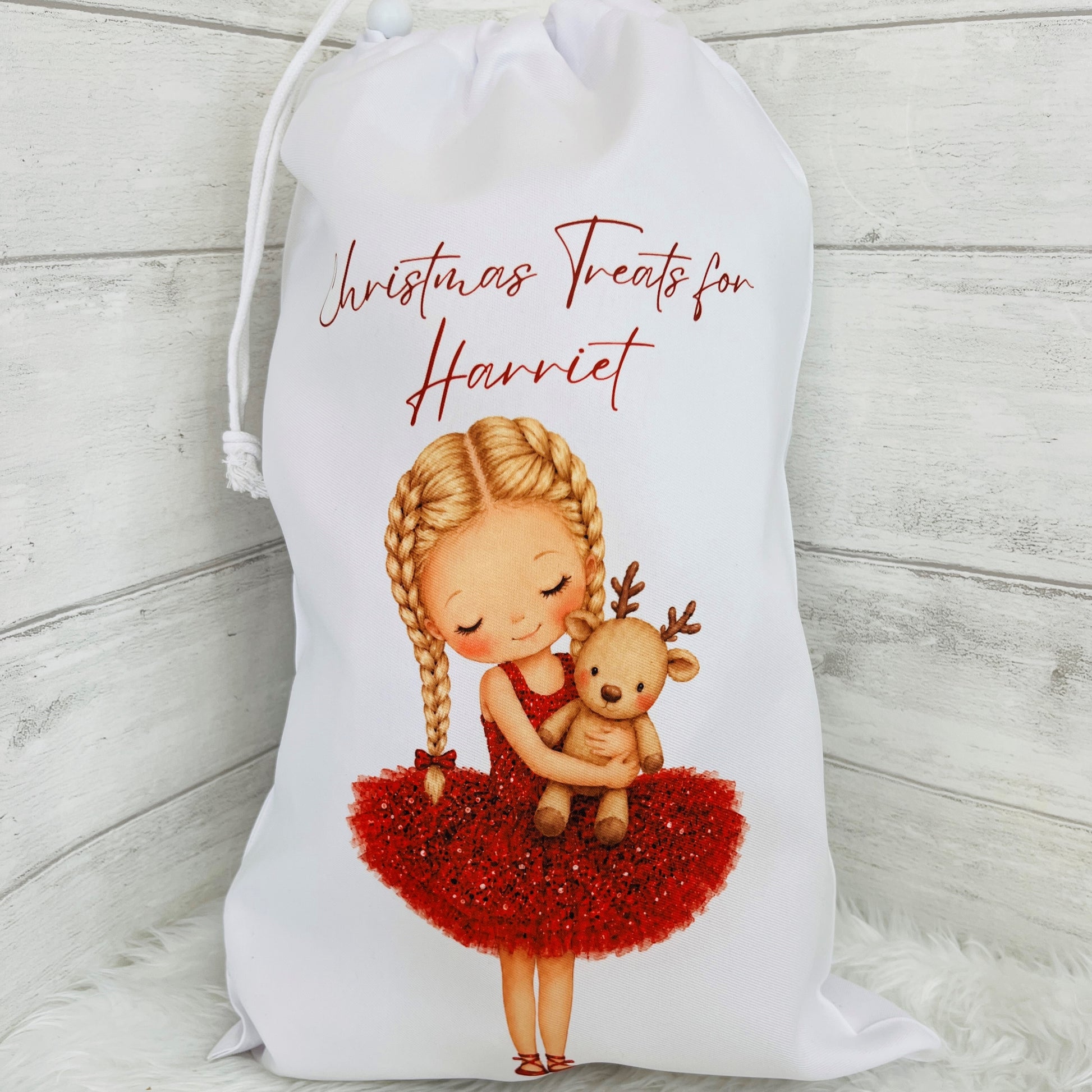 A white polyester gift bag with a drawstring closure, printed with the text 'Christmas Treats for Harriet' and an illustration of a blonde girl in a red dress hugging a teddy bear, set against a light wood plank background.