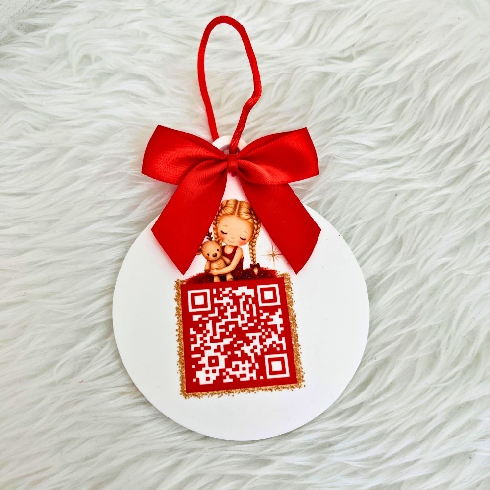 A round white acrylic santa tracker ornament with a red hanging ribbon bow, featuring a printed illustration of a girl holding a teddy bear above a red QR code design, photographed on a white faux-fur surface.