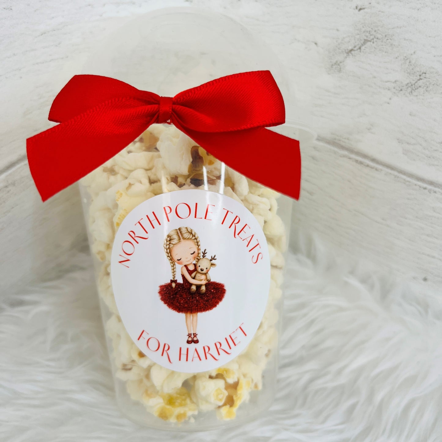 A clear plastic jar filled with popcorn, tied with a large red ribbon bow and a round white label that reads 'NORTH POLE TREATS FOR HARRIET,' set on a white faux-fur surface against a white wood background.