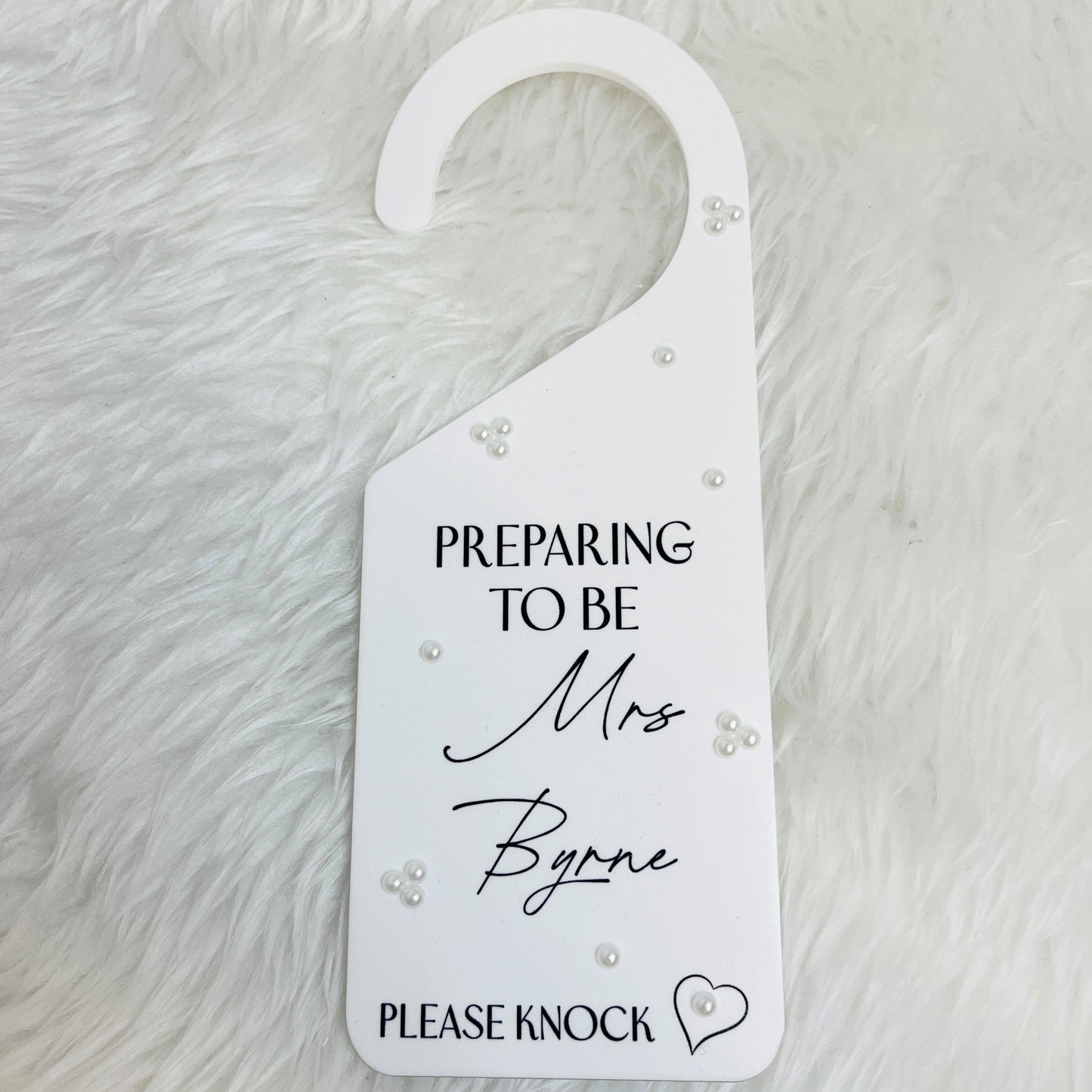 A white acrylic door hanger, printed with the text 'PREPARING TO BE Mrs Byrne PLEASE KNOCK' in black lettering, decorated with small white pearl-like beads, and placed on a white faux fur background.
