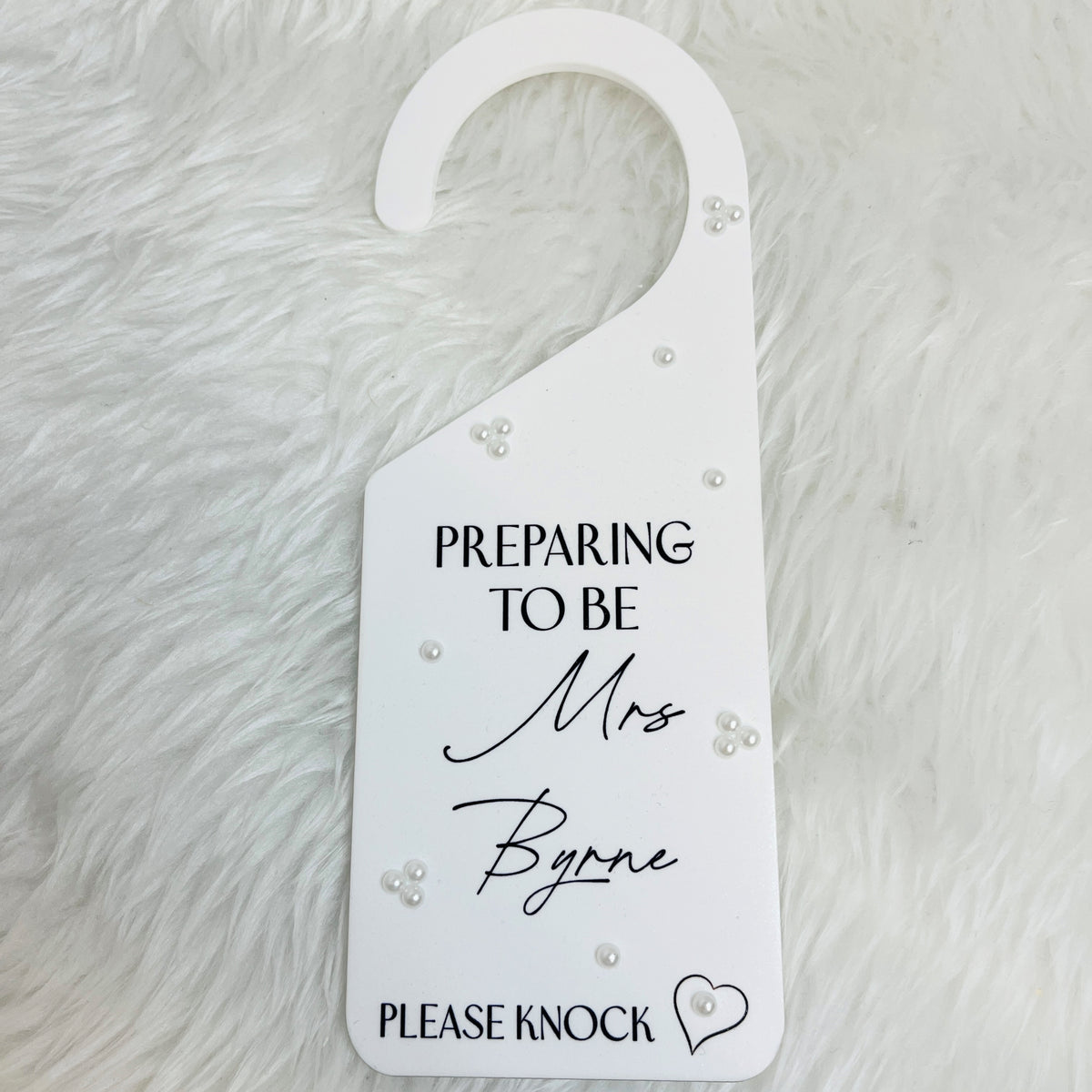 A white acrylic door hanger, printed with the text 'PREPARING TO BE Mrs Byrne PLEASE KNOCK' in black lettering, decorated with small white pearl-like beads, and placed on a white faux fur background.