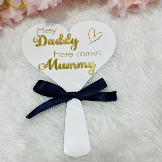 A white acrylic heart paddle with the text 'Hey Daddy here comes Mummy' in mirror gold acrylic with a navy bow on a white background