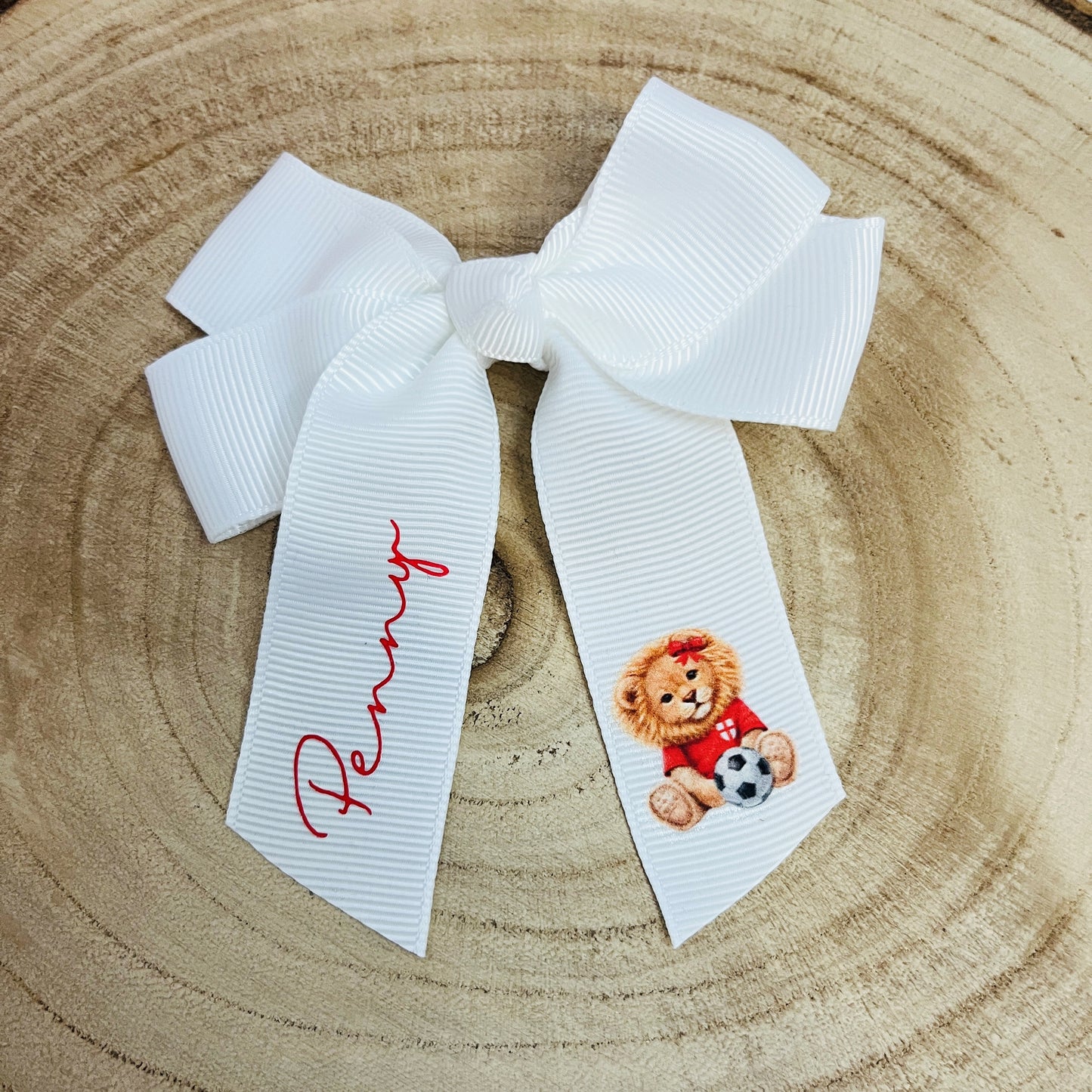 White 4inch grosgrain hair bow with a 'come on england' lion holding a football and the name 'Penny' on the other side on a wooden log slice background