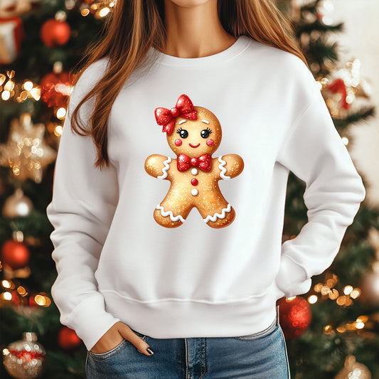 A woman wearing a white crewneck sweatshirt with a large, festive graphic of a smiling gingerbread man on the front. The gingerbread man is decorated with a red bow, red bow tie, and white icing details, set against a blurred Christmas tree background.