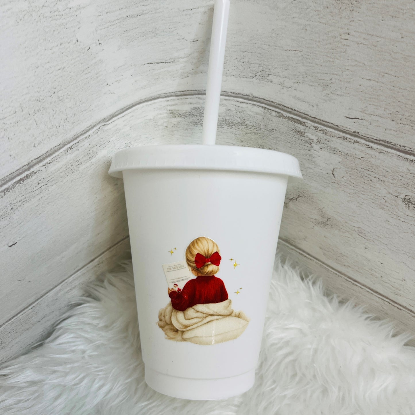 A white 16oz cold cup with a lid and straw, featuring a printed illustration of a girl in a red jumper and bow sitting on a blanket. The cup is placed on a white faux fur rug against a rustic white wood paneled background.