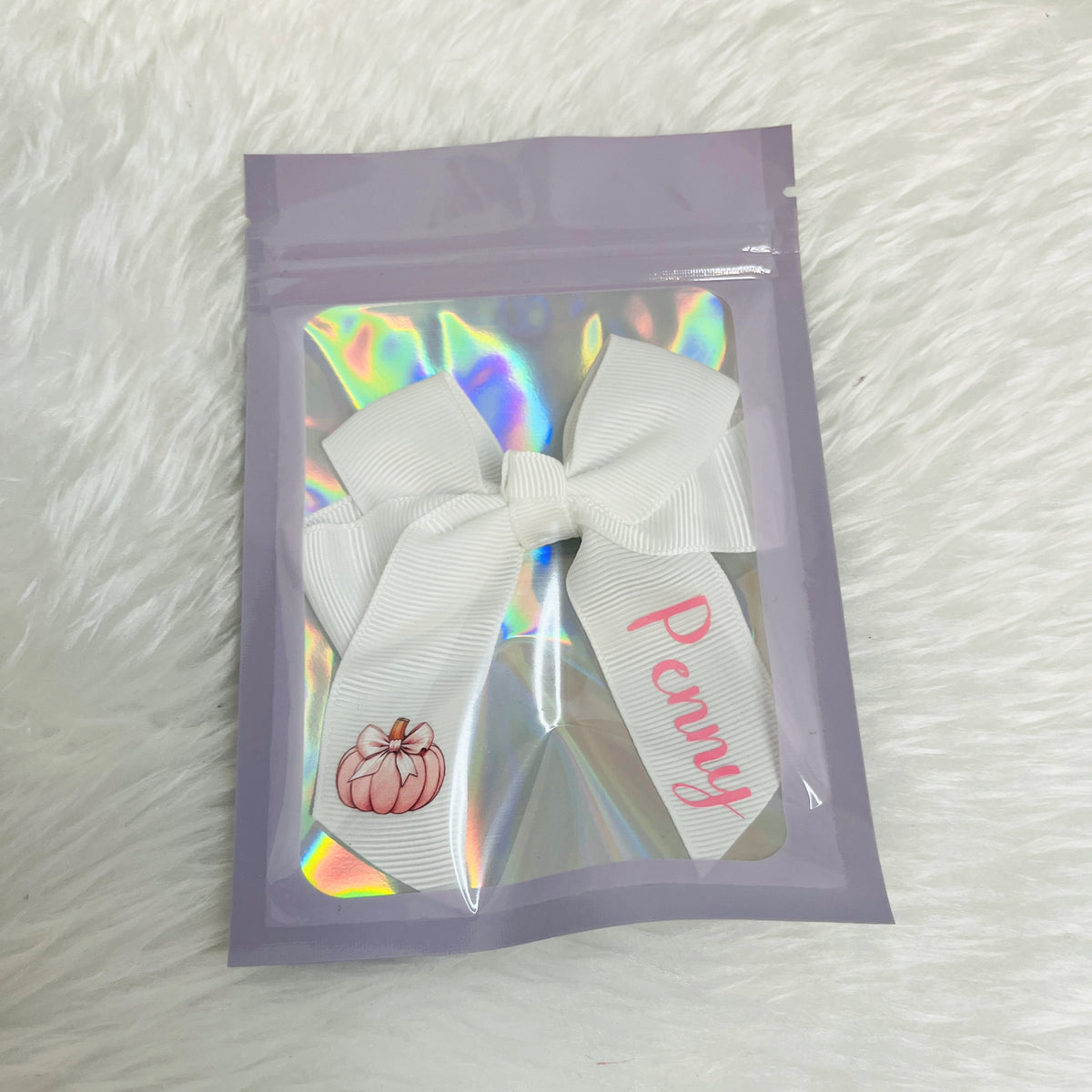 A white grosgrain ribbon bow with a pink pumpkin illustration and the name 'Penny' in pink script, attached to a clear windowed lavender pouch with an iridescent holographic insert, placed on a white faux fur background.