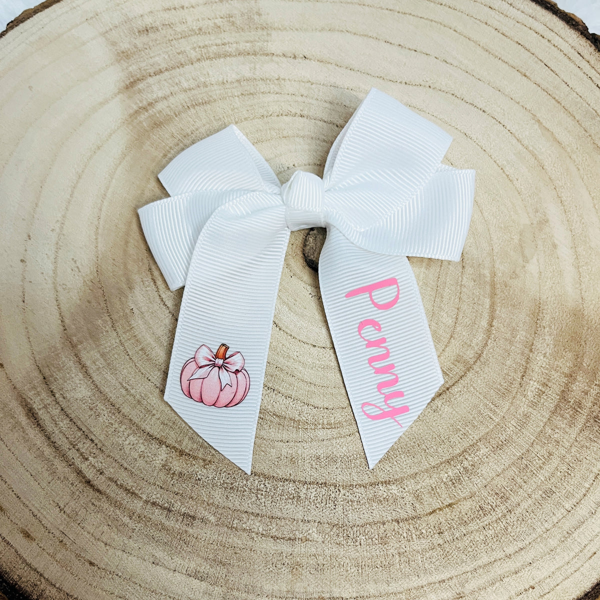 A white grosgrain ribbon hair bow with a pink pumpkin illustration on one tail and the name 'Penny' in pink script on the other, displayed on a circular wood slice background.
