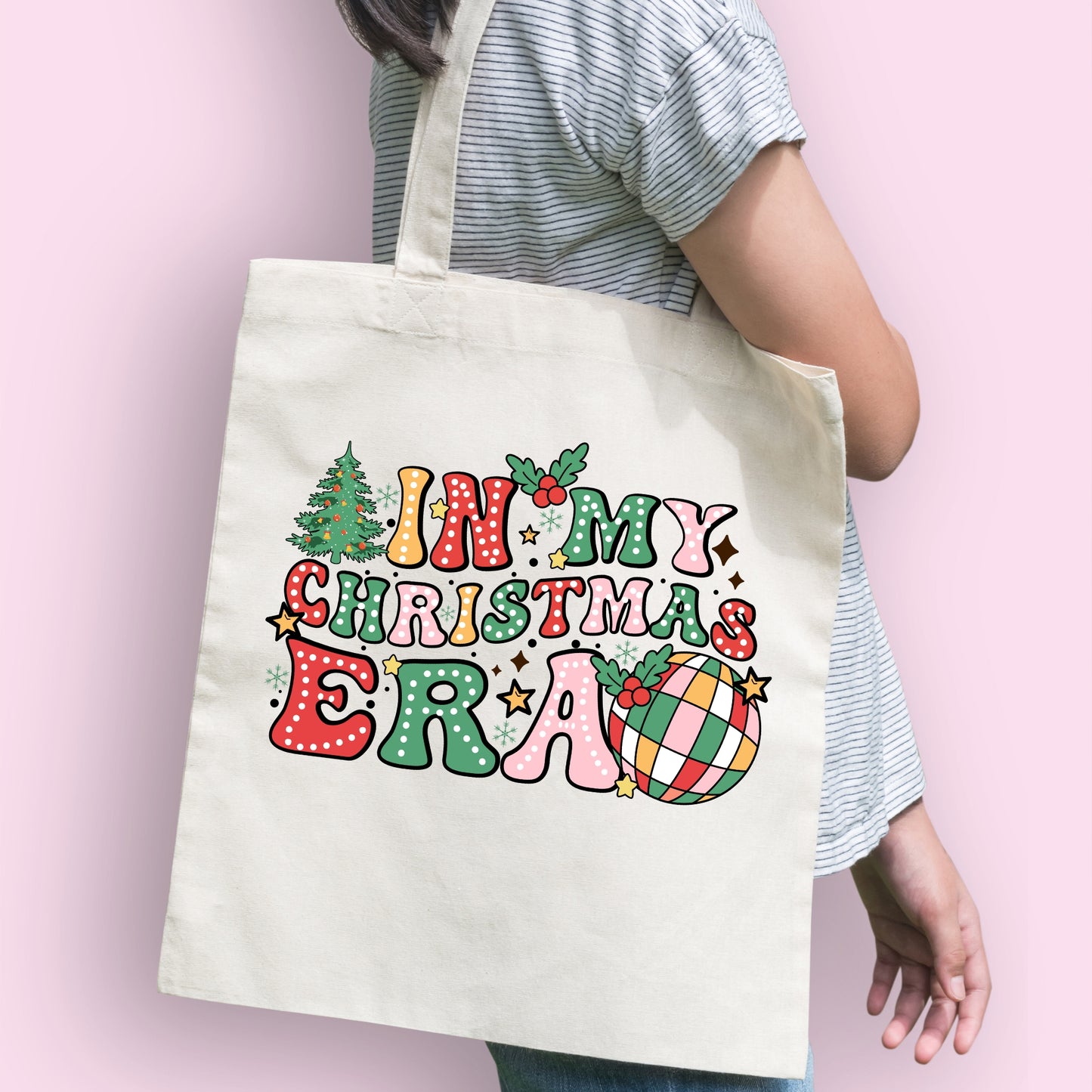 A model carrying a white tote bag with long handles and a 'in my christmas era' bright design