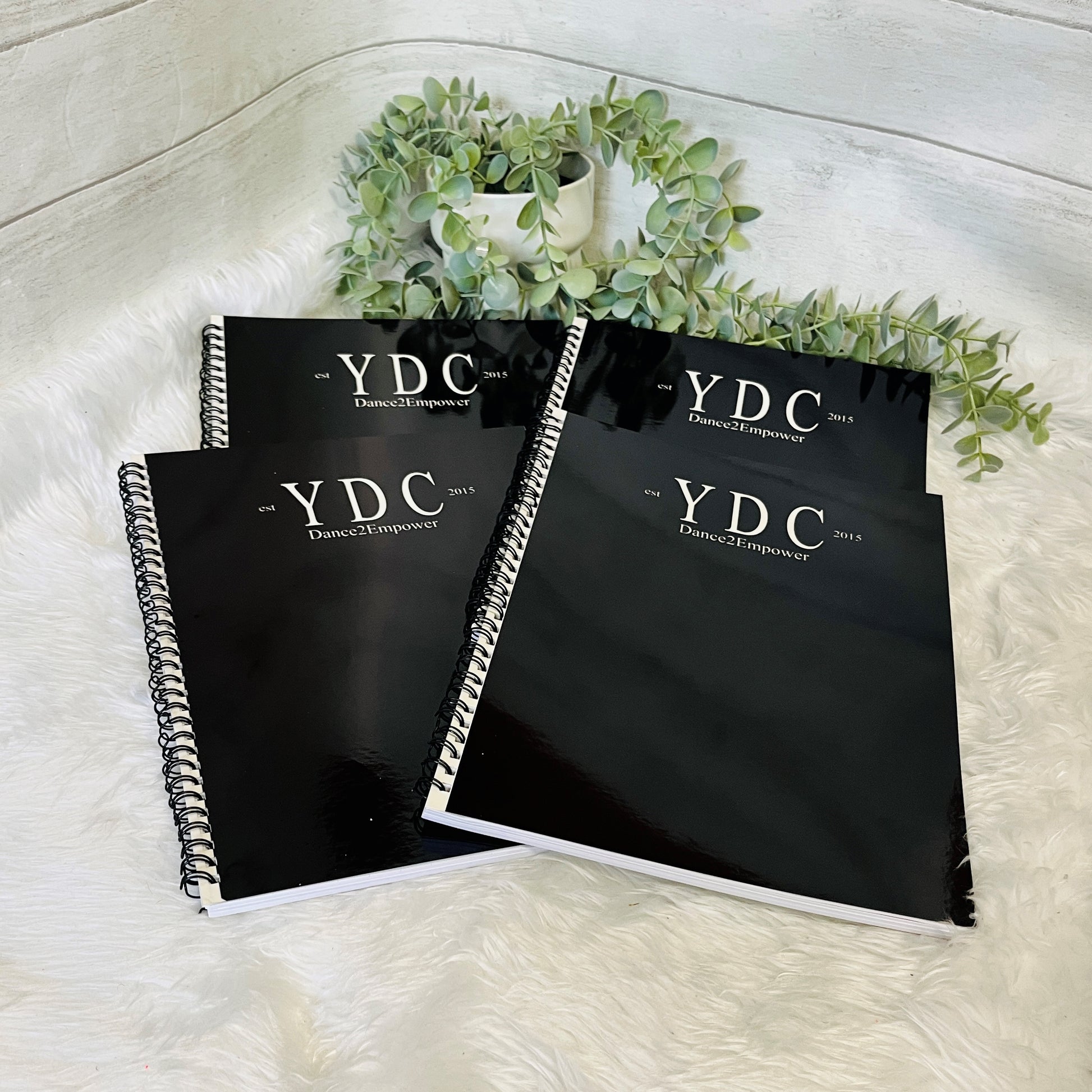 Three black notebooks with 'YDC Dance Empower' branding on a white surface with greenery.